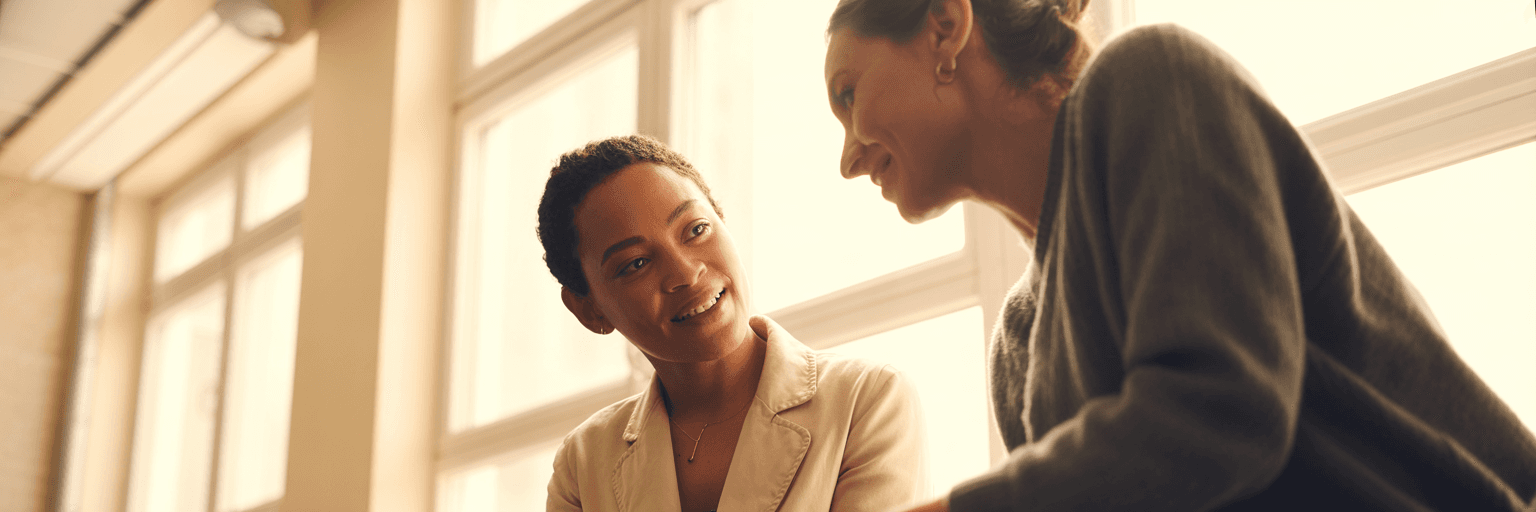 Two colleagues in a sunlit office looking together at something off camera, relaxed and collaborative.
