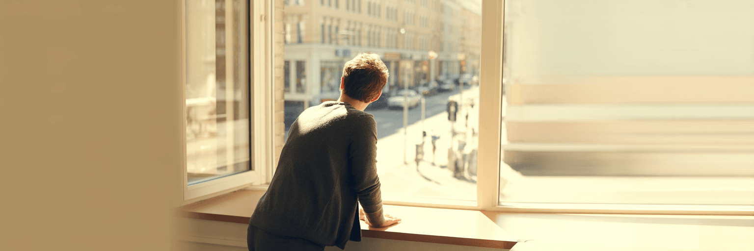 A person standing at a bright office window looking out over a sunlit street, seen from behind.